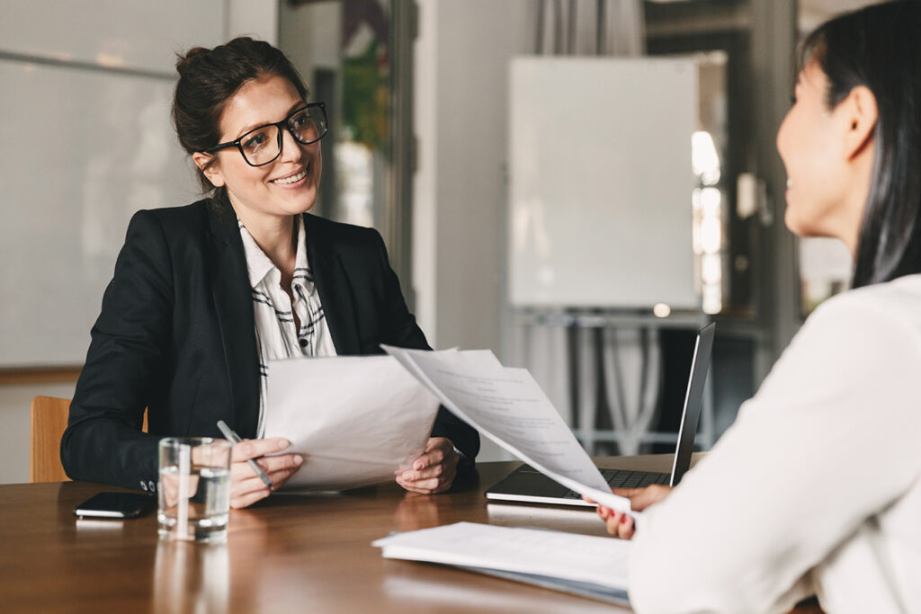 Portrait of smiling businesswoman holding resume and talking to female candidate during corporate meeting or job interview - business, career and placement concept