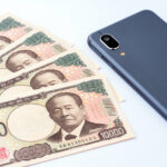 A 10,000 yen note and a smartphone. The Japanese inscription on the note reads "One Million Yen," "Bank of Japan Note," "Bank of Japan," "Produced by the National Printing Bureau," and "Shibusawa Eiic