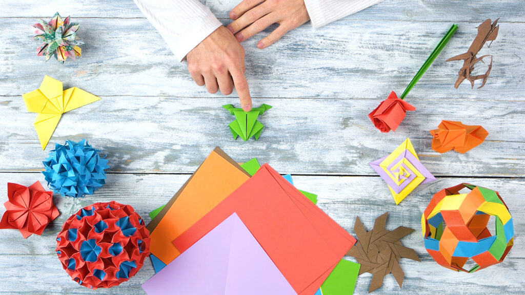 Man at origami folding lesson. Man pointing at origami frog with finger. Collection of beautiful origami figurines on wooden table. Traditional origami paper folding.